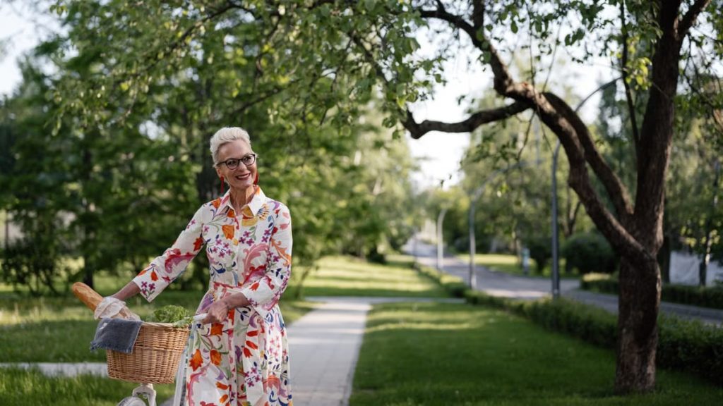 Smiling senior woman in floral dress walking bicycle outdoors on a sunny day.