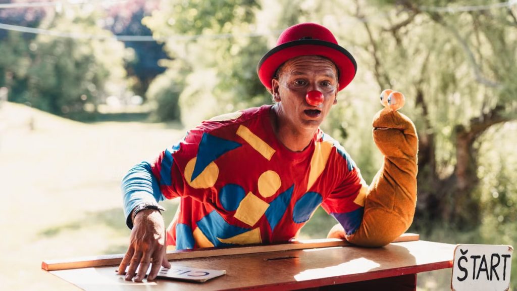 A clown wearing a colorful costume entertains with a puppet outdoors on a sunny day.